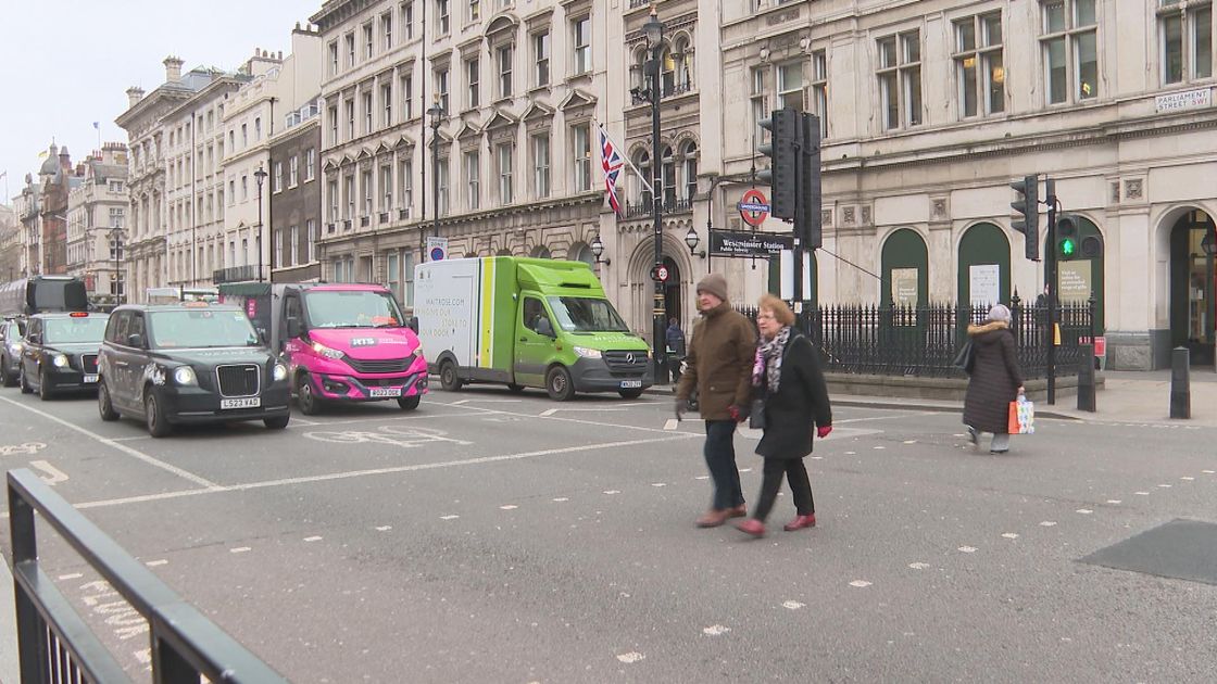 The image shows a street in Britain with parked cars, including electric vehicles, and pedestrians crossing in front of a historic building, highlighting the country's push for electric car subsidies and environmental policies.
